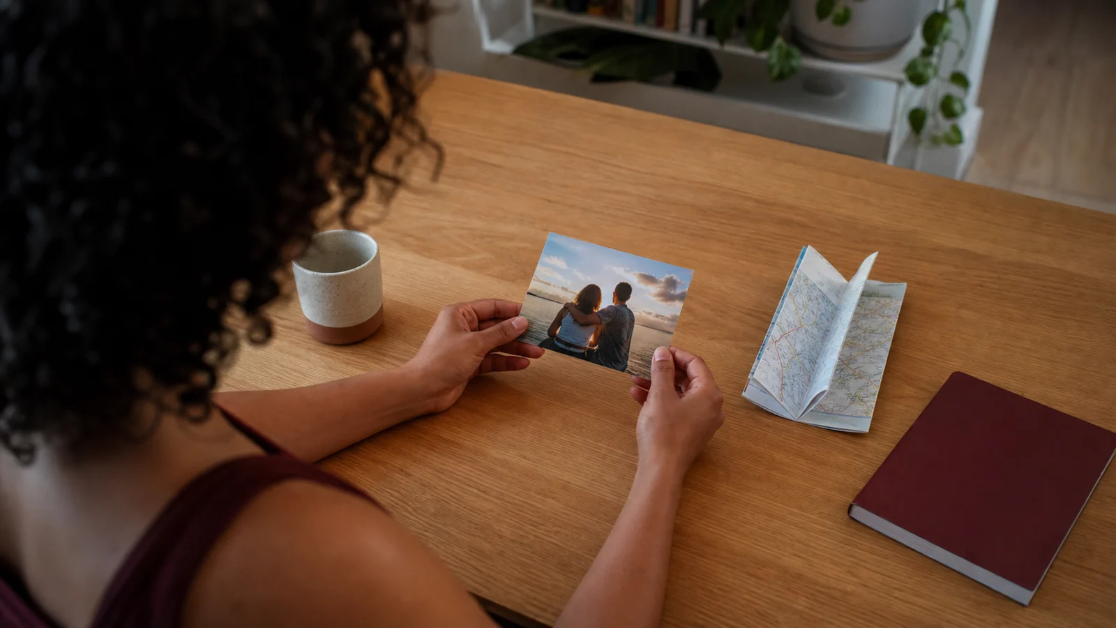 Pessoa segura uma fotografia diante de um mapa, caderno e objetos sobre a mesa